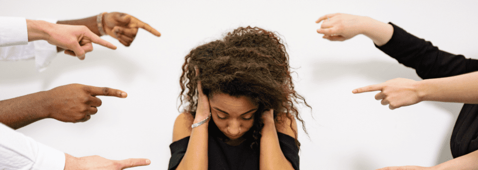 A young Black woman experiences harassment and discrimination. She stands against a white background with her hands over her ears.